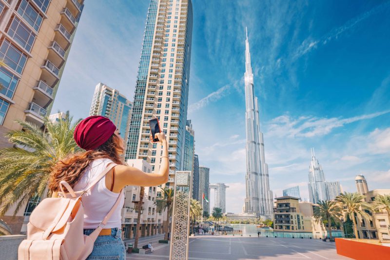 Burj Khalifa ve modern binalar arasında elinde telefonuyla fotoğraf çeken bir turist; unutulmaz anılarla dolu bir dubai şehir turu.