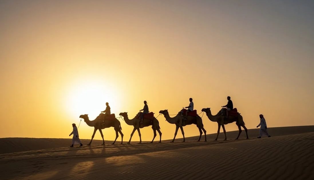 Dubai çölünde gün batımı sırasında kum tepesinin üzerinden geçen bir deve kervanı.